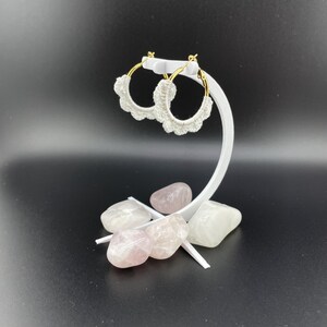 May include: White crocheted hoop earrings with gold metal hoops. The earrings are displayed on a white stand with pink and white stones.