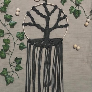 Op de afbeelding: Handgemaakte macramé wandhanger met een donkergrijs boomontwerp. De boom is geknoopt in een houten hoepel, met lange, bungelende franjes. Decoratief klimop en houten kralen omringen de wandhanger.