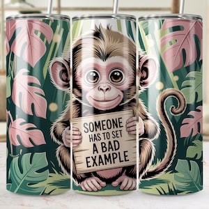 May include: A stainless steel tumbler featuring a cartoon monkey holding a sign that reads "SOMEONE HAS TO SET A BAD EXAMPLE." The design includes a jungle background with green and pink leaves. The tumbler has a straw.