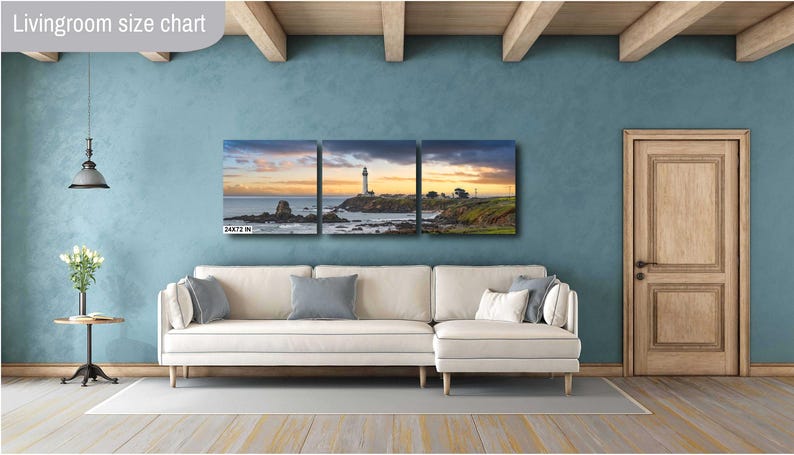 May include: A three-panel canvas print depicting a lighthouse on a rocky coastline. The print features a sunset sky with warm colors and a calm ocean. The print is displayed on a white couch in a living room setting.