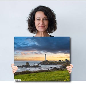 May include: A photograph of a lighthouse on a rocky coastline with a dramatic sky. The lighthouse is white and stands tall against a backdrop of dark clouds and a bright orange sunset. The ocean is calm and the water is a deep blue. The image is framed by a green grassy field.
