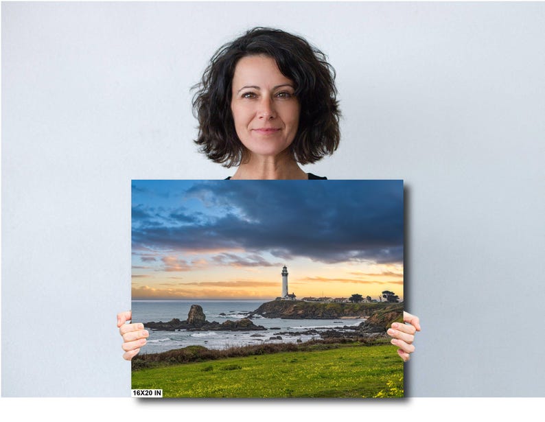 May include: A photograph of a lighthouse on a rocky coastline with a dramatic sky. The lighthouse is white and stands tall against a backdrop of dark clouds and a bright orange sunset. The ocean is calm and the water is a deep blue. The image is framed by a green grassy field.