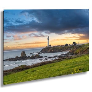 May include: A white lighthouse stands tall on a rocky cliff overlooking a vast ocean. The sky is a mix of blue, gray, and orange clouds, creating a dramatic sunset scene. The foreground features a grassy field with wildflowers.