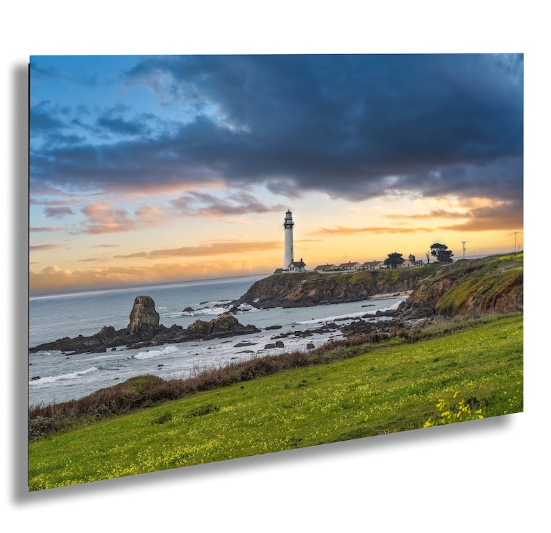 May include: A white lighthouse stands tall on a rocky cliff overlooking a vast ocean. The sky is a mix of blue, gray, and orange clouds, creating a dramatic sunset scene. The foreground features a grassy field with wildflowers.