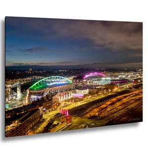 May include: A nighttime cityscape featuring illuminated stadiums and buildings under a twilight sky. The stadiums are lit with vibrant green, blue, and pink lights, creating a colorful contrast against the dark sky. The city lights twinkle in the distance.