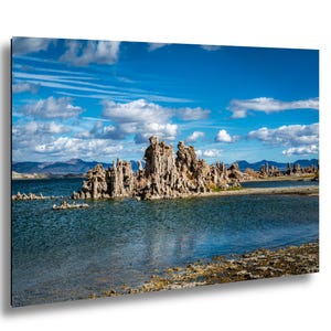 May include: A scenic landscape photograph featuring a lake with unique rock formations. The water is a deep blue, reflecting the sky with fluffy white clouds. The rock formations are a light brown color, contrasting with the blue water and sky.