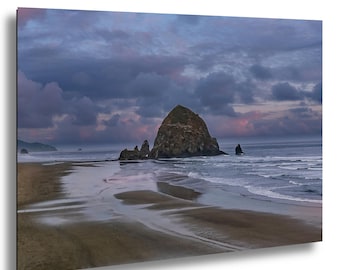 May include: A scenic landscape photograph of a beach at dusk. The image features a large rock formation in the ocean, with waves gently lapping the shore. The sky is filled with dramatic, dark clouds.