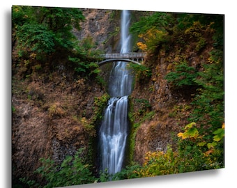 May include: A scenic photograph of a waterfall cascading down a rocky cliff face. A small stone bridge arches over the falls, surrounded by lush green and autumn-colored foliage. The image captures the natural beauty of the landscape.