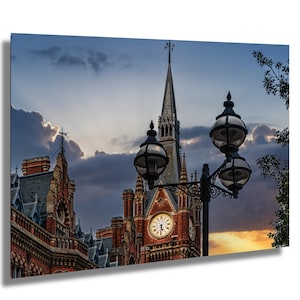 May include: A red brick building with a tall clock tower and a street lamp in the foreground. The clock tower has a clock face with Roman numerals and a spire. The sky is a mix of blue, gray, and orange.