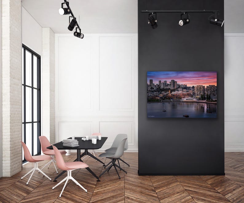 a modern, minimalist living room with a large black wall as the focal point. On the wall, there is a framed image of a city skyline at sunset. The room is furnished with a black dining table surrounded by four chairs, and a flat-screen TV is mounted on the wall. The overall design and color scheme of the room create a sleek and sophisticated atmosphere.