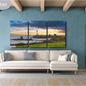 May include: A three-panel canvas print depicting a lighthouse on a rocky coastline with a sunset sky. The print is displayed on a white sectional sofa in a living room setting.