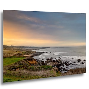 May include: Coastal landscape print featuring a dramatic sky with orange and blue hues. The image showcases a rocky shoreline with waves crashing against the rocks, and a grassy area in the foreground. The overall theme is nature and landscape art.