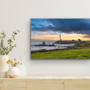 May include: A white lighthouse stands on a rocky outcropping overlooking a blue ocean with a dramatic cloudy sky. The sun is setting, casting a warm glow on the horizon.