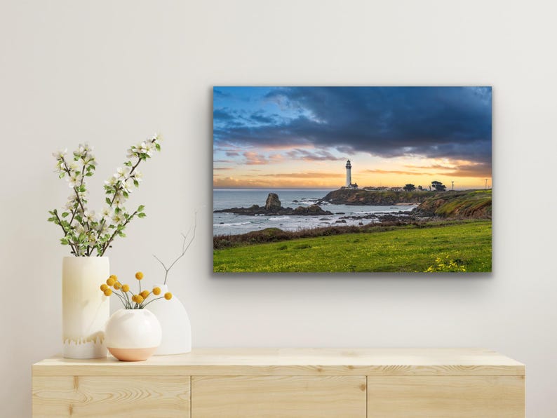 May include: A white lighthouse stands on a rocky outcropping overlooking a blue ocean with a dramatic cloudy sky. The sun is setting, casting a warm glow on the horizon.