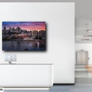 a modern office space with a large wall-mounted painting depicting a cityscape at sunset. The painting is framed in white and hangs on a white wall. The office space has a long hallway with glass doors and a white desk with a computer monitor.