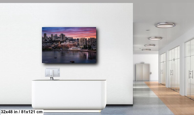 a modern office space with a large wall-mounted painting depicting a cityscape at sunset. The painting is framed in white and hangs on a white wall. The office space has a long hallway with glass doors and a white desk with a computer monitor.