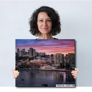 A woman holding a large framed print of a cityscape at sunset, with the city skyline and water visible in the image.