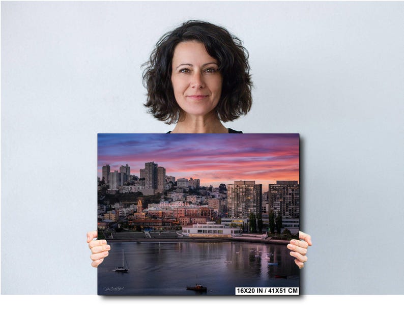 A woman holding a large framed print of a cityscape at sunset, with the city skyline and water visible in the image.