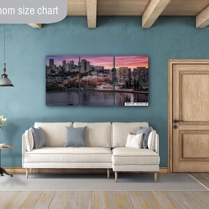 a living room with a large, colorful cityscape painting hanging on the wall. The painting features a skyline with tall buildings and a body of water in the foreground. The room has a white sofa, a wooden door, and a small table with a vase of flowers.
