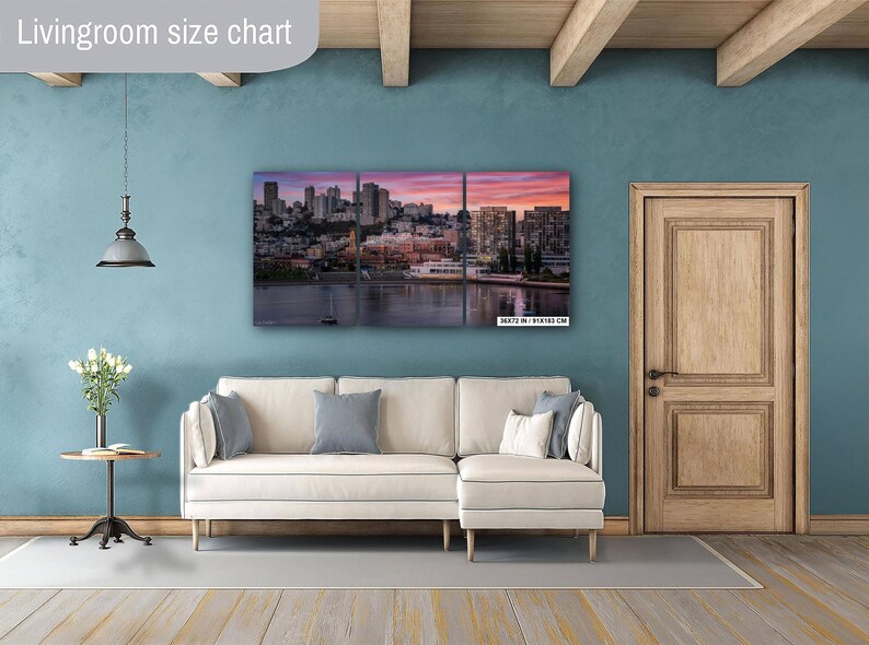 a living room with a large, colorful cityscape painting hanging on the wall. The painting features a skyline with tall buildings and a body of water in the foreground. The room has a white sofa, a wooden door, and a small table with a vase of flowers.