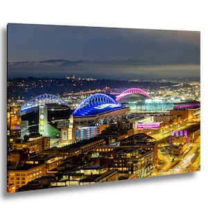 May include: A nighttime cityscape featuring illuminated stadiums and buildings. The stadiums are lit with blue and pink lights, and the city skyline is visible in the background. The image is a vibrant display of urban architecture.