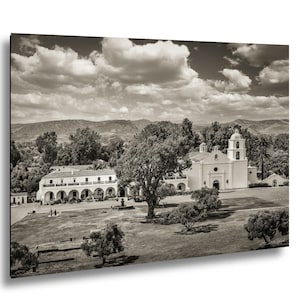 May include: Sepia-toned print of a historic mission with a white facade, arched walkways, and a bell tower. The building is surrounded by trees and a grassy lawn, with mountains and a cloudy sky in the background.