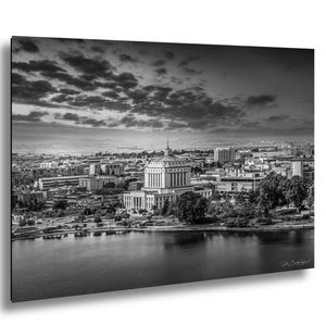 May include: Black and white photograph of a cityscape featuring a large, ornate building with a domed roof. The building is situated near a body of water, with other buildings and a cloudy sky in the background. The image is a landscape orientation.