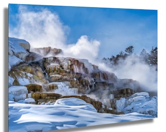 Könnte beinhalten: Landschaftsfotografie einer geothermischen heißen Quelle im Winter. Das Bild zeigt eine kaskadenartige, geschichtete Felsformation, die mit Schnee und Eis bedeckt ist, wobei Dampf in einen hellblauen Himmel aufsteigt. Die Felsen haben Braun- und Orangetöne.