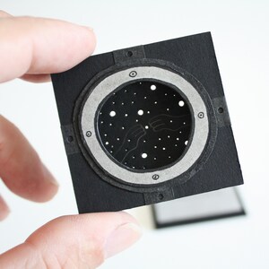 May include: A black paper craft with a circular cutout featuring a starry night sky with two hands reaching out to each other. The cutout is surrounded by a silver ring with a black border.