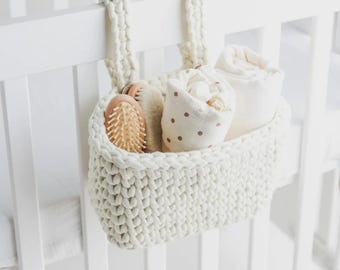 May include: A cream-colored, hand-knitted basket hangs from a white crib. The basket contains a wooden hairbrush, a soft brush, a polka-dot swaddling blanket, and a rolled-up white blanket. The basket is designed for baby essentials.