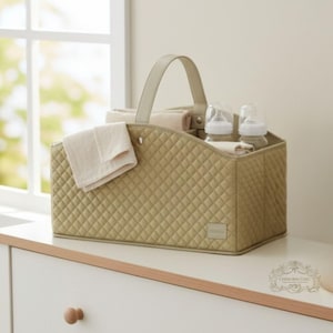 Op de afbeelding: Een gewatteerde, olijfgroene babycaddy met een handvat, met twee babyflessen, een gevouwen doek en opgerolde handdoeken. De caddy staat op een wit oppervlak bij een raam. De caddy heeft een ruitpatroon.