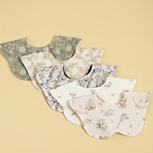 May include: A collection of six baby bibs with various floral and toy patterns. The bibs feature a rounded shape with a curved neckline. Colors include green, beige, blue, and brown on a white or cream background.