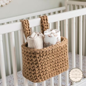 May include: A brown, hand-knitted basket hanging on a white crib rail. The basket contains two rolled-up baby blankets. The basket is made of thick yarn and has two handles for hanging. The blankets are light-colored with floral patterns.