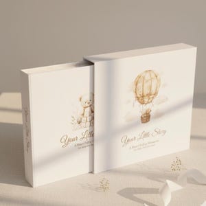 May include: White baby memory book with a hot air balloon and teddy bear illustration. The book cover reads "Your Little Story" in elegant script. The book is designed to hold baby photos and memories.