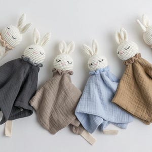 Può includere: Una collezione di copertine di sicurezza a forma di coniglietto all'uncinetto in vari colori. Ogni coniglietto ha una testa bianca all'uncinetto con gli occhi chiusi e lunghe orecchie. Le coperte sono in tonalità di grigio, beige e blu, con un bordo a balze intorno al collo.