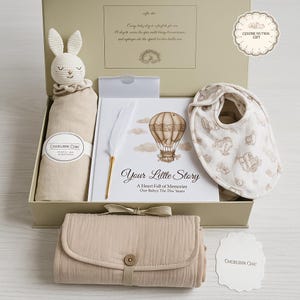 May include: A beige baby gift set in a box. The set includes a bunny plush, a swaddle blanket, a bib, a changing pad, and a memory book with a feather pen. The book cover reads "Your Little Story".