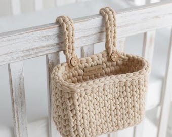 May include: A beige, hand-crocheted basket with two looped handles, attached to a white crib rail. The basket has a rectangular shape and is secured with wooden buttons. The label reads "HEAVEN CHIC".