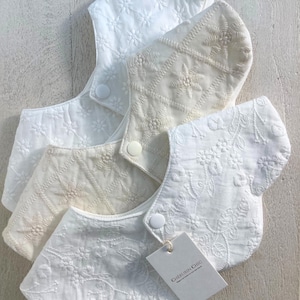 Op de afbeelding: Vier witte en crèmekleurige baby slabbetjes met ingewikkelde bloemenborduursels. Elk slabbetje heeft een drukknoopsluiting en een geschulpte rand. Een label met de tekst "CHERUBIN CHIC" is bevestigd.