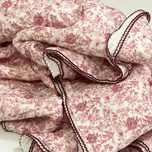 Op de afbeelding: Een gevouwen deken met een bloemenpatroon in wit en stoffig roze. De deken heeft een herhalend patroon van rozen en bladeren. De randen zijn afgewerkt met een witte rand en een donkerrode zigzagsteek.