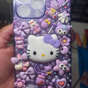 Puede incluir: Una funda transparente de teléfono de color morado con un diseño de Hello Kitty. La funda está decorada con varios abalorios, incluyendo lazos, estrellas, osos y caramelos. La funda está cubierta de purpurina.