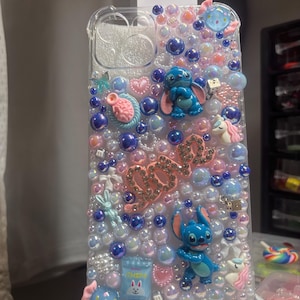 May include: A clear phone case decorated with blue and pink iridescent beads, rhinestones, and charms. The case features two blue Stitch characters, a pink heart, a unicorn, and a bunny. The word "Love" is spelled out in rhinestones.