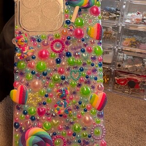Könnte beinhalten: Transparente Handyhülle, verziert mit bunten Perlen, Lollis und herzförmigen Akzenten. Die Hülle weist eine Vielzahl von Farben auf, darunter Rosa, Grün, Blau und Regenbogenfarben, wodurch ein verspieltes und lebendiges Design entsteht.