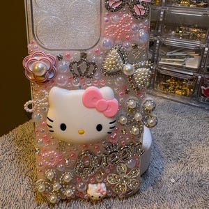 Può includere: Una custodia trasparente per telefono decorata con abbellimenti rosa, bianchi e argentati. Presenta un design di Hello Kitty, una farfalla, un fiocco e la parola "Queen". La custodia è adornata con perle, strass e accenti floreali.