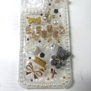 Op de afbeelding: Een transparante telefoonhoesje versierd met witte parels, gouden en zilveren versieringen, een zwarte beer, een vlinder, een kruis, een strik en het woord "BLESSED".