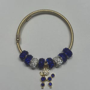 Puede incluir: Pulsera dorada con cuentas de pedrería azul y plateada. Un charm con forma de caniche, adornado con pedrería azul y transparente, cuelga de la pulsera. La pulsera está sobre una superficie gris claro.