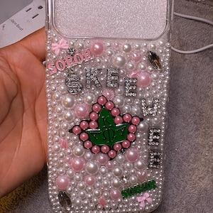 Puede incluir: Funda de teléfono transparente adornada con perlas blancas y rosas, y pedrería. La funda presenta las palabras "SOPOR" y "SKEE WEE" en letras de pedrería, junto con un emblema verde y rosa.