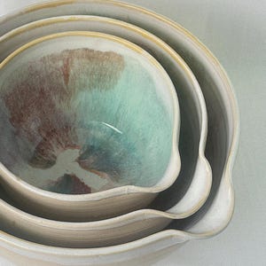 May include: A set of three ceramic mixing bowls nested together. The bowls have a light beige exterior with a yellow rim. The interior of the bowls have a turquoise and brown glaze.