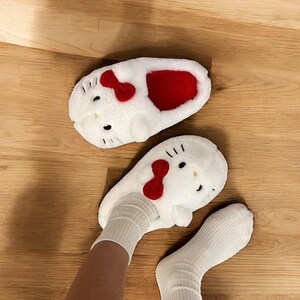 May include: White fuzzy slippers with a Hello Kitty design. The slippers have red bows and a red interior. The slippers are being worn by a person with white socks.