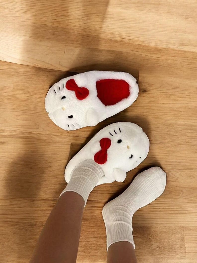 May include: White fuzzy slippers with a Hello Kitty design. The slippers have red bows and a red interior. The slippers are being worn by a person with white socks.
