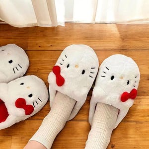 May include: White fuzzy slippers with a red bow and a Hello Kitty face on the front. The slippers are worn on a person's feet.